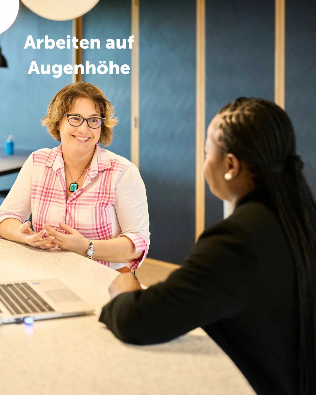 Zwei Kolleginnen sitzen an einem Tisch und lächeln sich an.