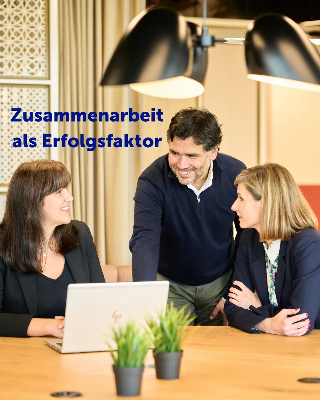 Zwei Kolleginnen sitzen an einem Tisch vor einem aufgeklappten Laptop. Sie wenden sich einem Kollegen zu, der sich zu ihnen leicht herunterbeugt. 