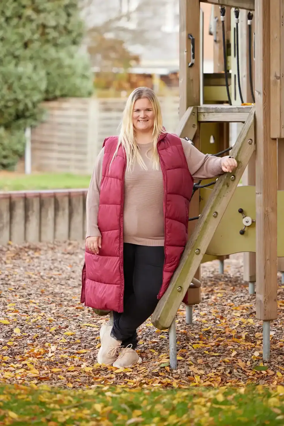 Frau in pinker Daunenweste steht vor einem Spielgerät auf einem Spielplatz.