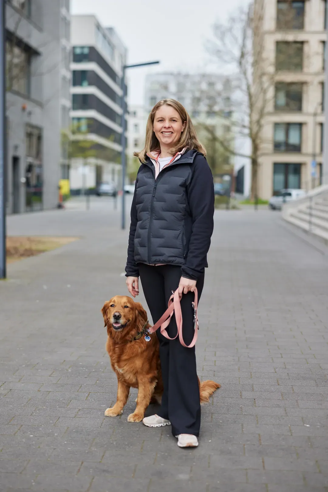 Frau mit dunkelblauer Jacke steht mit einem Hund auf einem Gehweg in der Stadt.