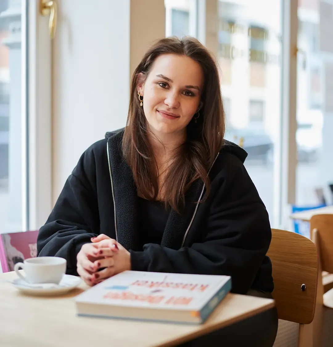 Junge Frau sitzt entspannt an einem Tisch mit einem Buch und einer Tasse Kaffee.