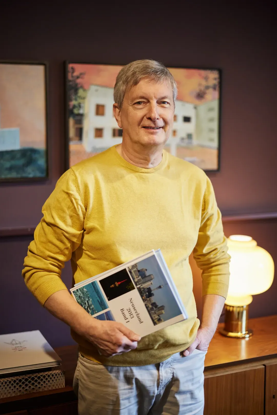 Mann in gelbem Pullover hält ein Buch vor seinem Oberkörper in einem indirekt beleuchteten Wohnraum.